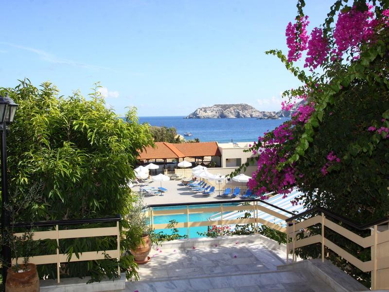 Vue sur une zone de piscine avec chaises longues, plantes en fleurs et mer en arrière-plan.
