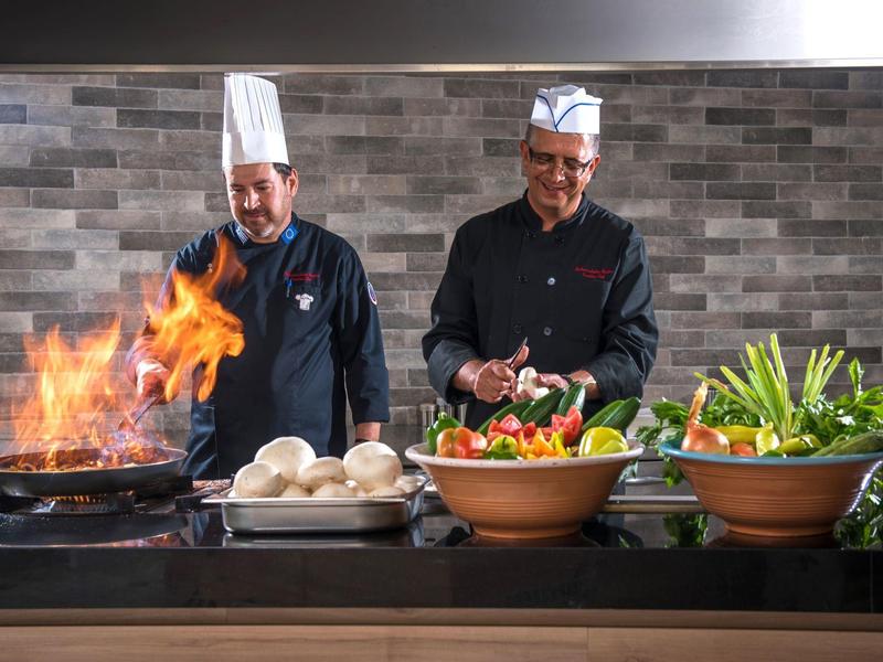 Deux chefs préparent des légumes frais et un plat enflammé dans une cuisine d'hôtel.