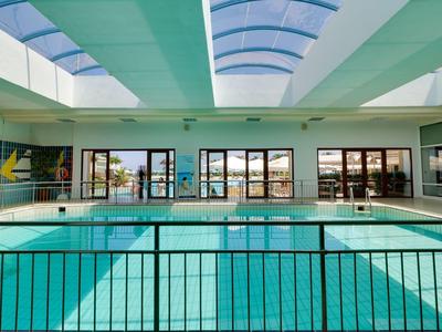 Innenraum mit hellem Schwimmbad, großen Fenstern und einem gebogenen Glasdach.