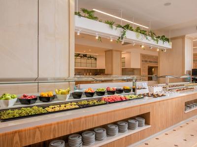 Área de buffet con frutas frescas, verduras y pan en un restaurante de hotel moderno y luminoso.