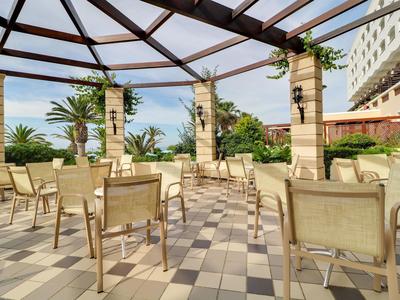 Terraza al aire libre con sillas y mesas bajo una estructura acristalada junto a un hotel.