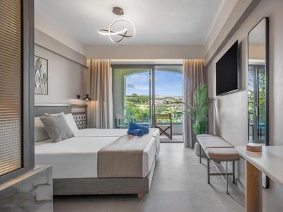 Modern eingerichtetes Hotelzimmer mit Doppelbett, TV, Balkon und Blick auf grüne Hügel.