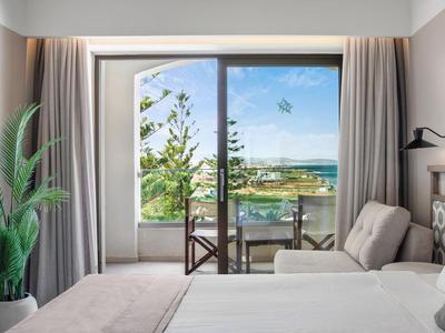 Hotelzimmer mit Bett, Sessel und Balkon mit Meerblick, umgeben von grünen Bäumen.