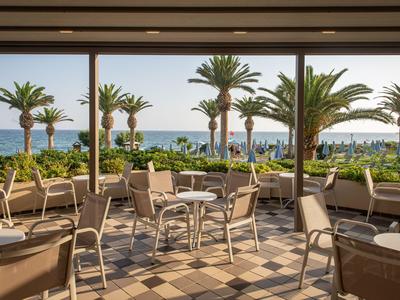 Open terras met tafels en stoelen, uitzicht op zee en palmbomen.