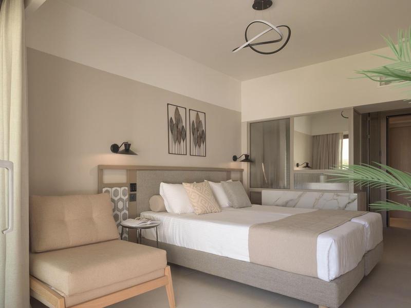 Habitación de hotel luminosa con cama grande, sillón, arte moderno y pared con espejo.