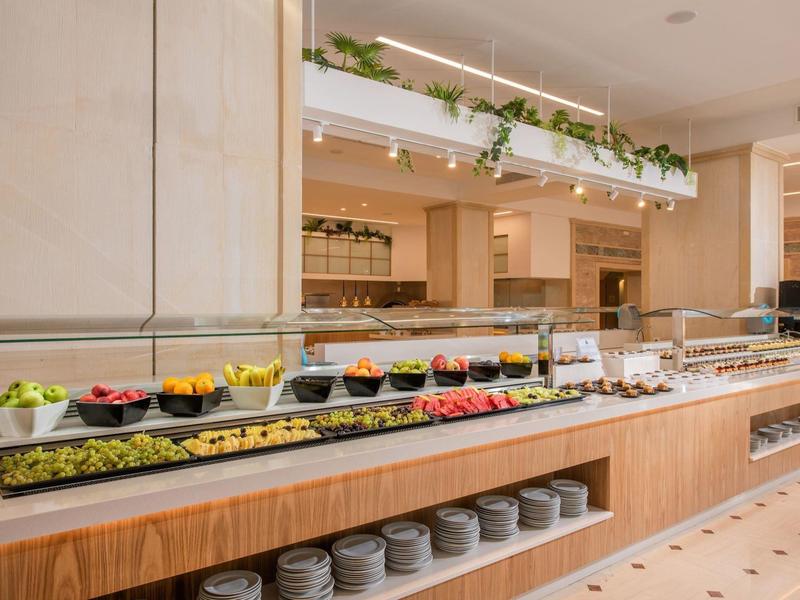 Área de buffet con frutas frescas, verduras y pan en un restaurante de hotel moderno y luminoso.