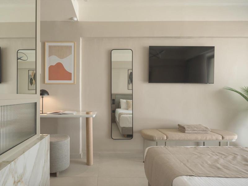 Habitación de hotel moderna con cama, espejo de pared, televisor y decoración minimalista en tonos neutros.