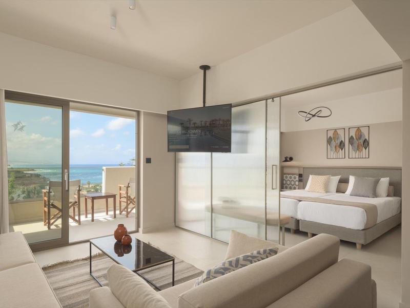 Habitación de hotel moderna con sofá, televisor de pantalla plana y vista al mar a través de grandes ventanas y balcón.