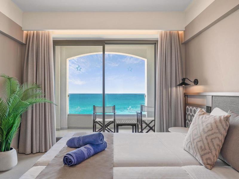 Habitación de hotel con vista al mar, cama, mesa con sillas y ambiente relajado en la playa.