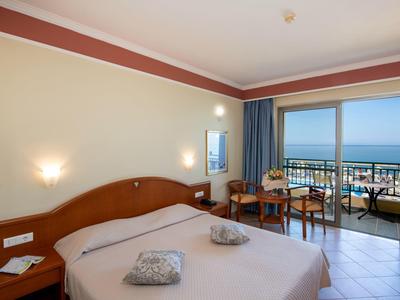 Helles Schlafzimmer mit Doppelbett, Balkon, Meerblick, Vorhängen und kleinen Holzmöbeln.