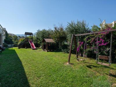 Garten mit grünem Rasen, Schaukel, Sandkasten, Rutsche und Bäumen im Hintergrund.
