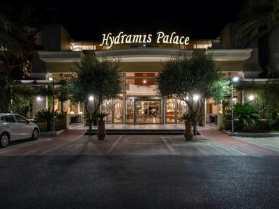 Eingang zum Hydramis Palace bei Nacht mit beleuchteten Bäumen und parkenden Autos.