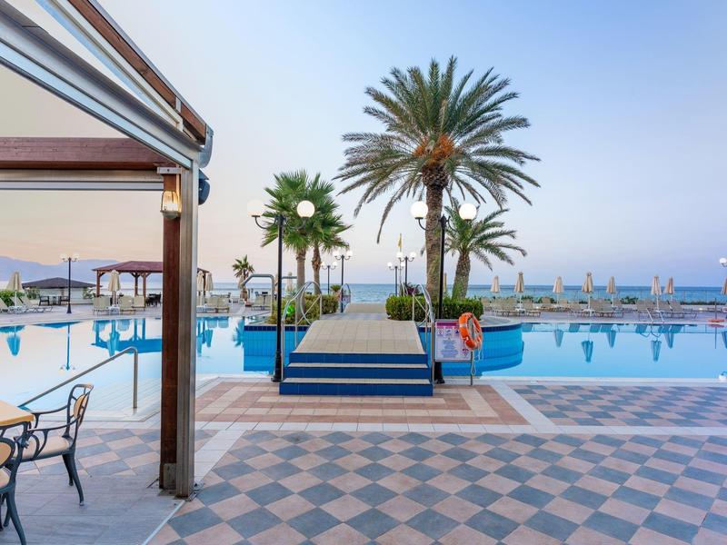 Großer Poolbereich mit Palmen, blauer Wasserfläche, Stühlen und Meer im Hintergrund bei Abendlicht.