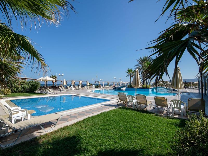 Blick auf einen blauen Pool mit weißen Liegestühlen, Palmen und blauem Himmel am Meer.