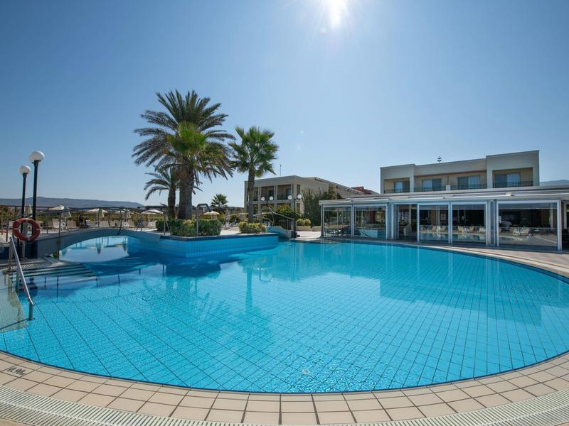 Großer runder Pool mit klar blauem Wasser, Palmen und weißem Gebäude bei sonnigem Himmel.