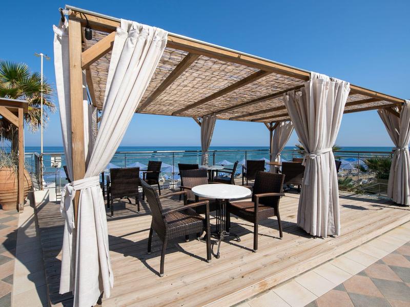 Offene Pergola mit weißen Vorhängen, schwarzen Stühlen und Tischen mit Meerblick bei Sonnenschein.
