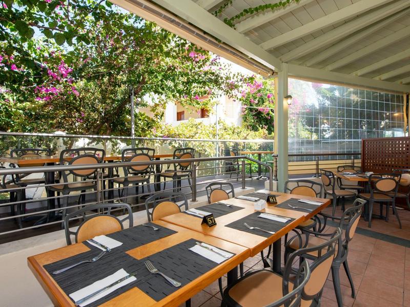 Offener Essbereich mit Holztischen, Metallstühlen und grüner Pflanzenkulisse im Freien.