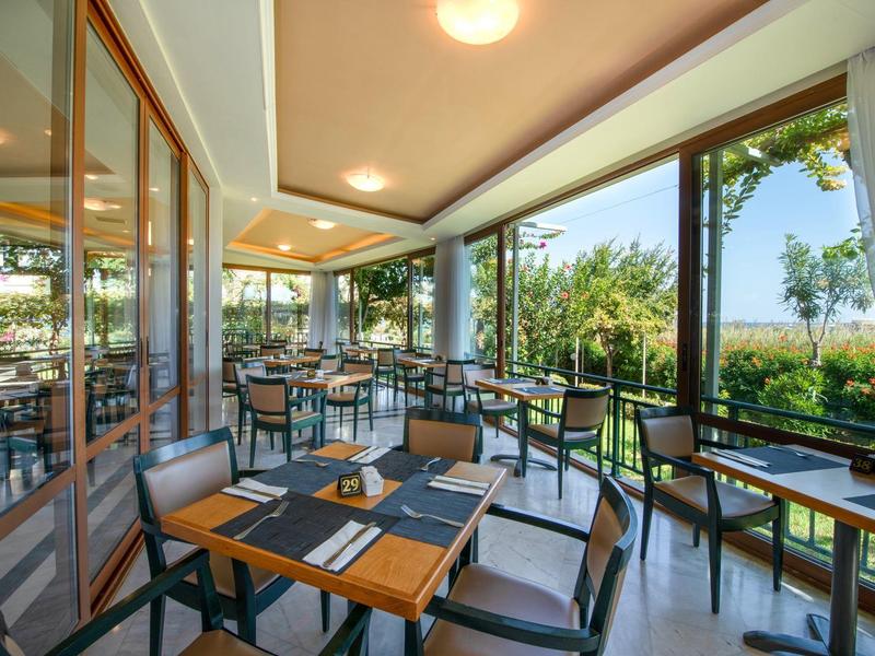 Moderner, heller Wintergarten mit Holztischen, schwarzen Stühlen und großer Glasfront zur grünen Außenansicht.