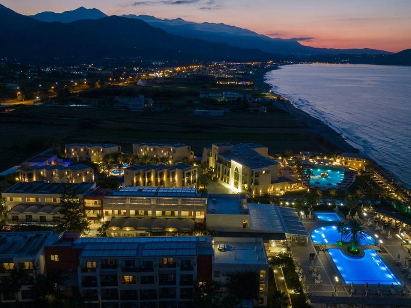 Lichtbeleuchtetes Hotel und Poolanlage an Küste bei Sonnenuntergang mit Bergsilhouette im Hintergrund.