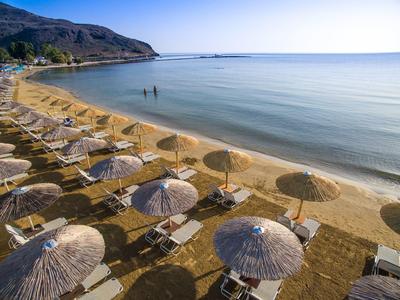 Słoneczna plaża z trzcinowymi parasolami i leżakami nad spokojną, czystą wodą z górami w tle.