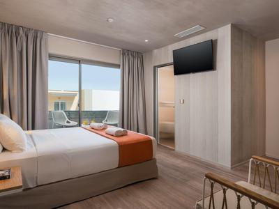 Nowoczesny pokój hotelowy z podwójnym łóżkiem, telewizorem, balkonem i neutralnym wystrojem z pomarańczowymi akcentami.