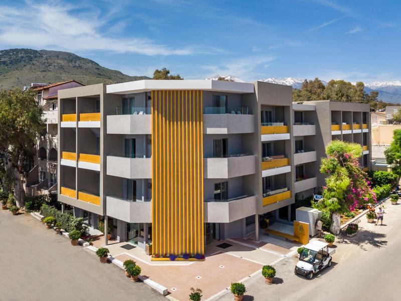 Nowoczesny budynek hotelu z żółtymi akcentami, otoczony zielenią i widokiem na góry.