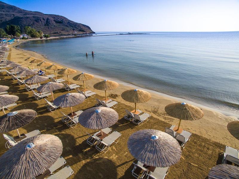 Słoneczna plaża z trzcinowymi parasolami i leżakami nad spokojną, czystą wodą z górami w tle.