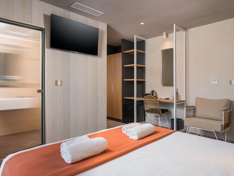 Nowoczesny pokój hotelowy z łóżkiem, pomarańczowym kocem, ręcznikami, biurkiem, krzesłem i telewizorem.