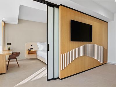 Modernes Hotelzimmer mit Holzwand, TV, Arbeitsbereich und großer Glasschiebetür zum Schlafzimmer.