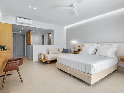 Habitación de hotel moderna con cama doble grande, sofá, escritorio y diseño minimalista en tonos claros.