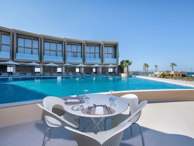 Modern hotel complex with large pool and outdoor table overlooking the sea.