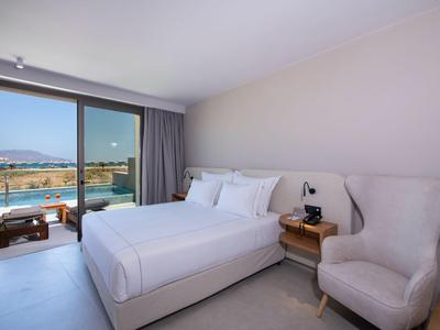Habitación de hotel moderna con cama doble, sillón y vista a la piscina y el paisaje.