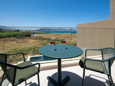 Balcón con mesa y dos sillas con vista a jardín y agua bajo cielo despejado.