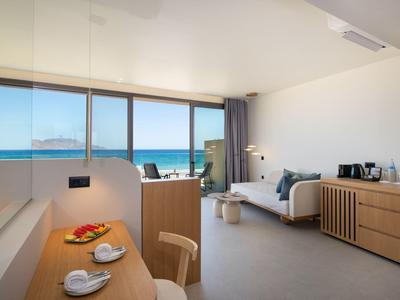 Habitación de hotel luminosa con vista al mar, decoración moderna y mesa con cesta de frutas.