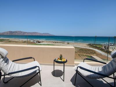 Balcón con dos sillas, una mesa y vista despejada al mar y la playa bajo un cielo azul.