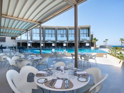 Área exterior moderna de hotel con piscina, tumbonas y mesas preparadas bajo un cielo azul.