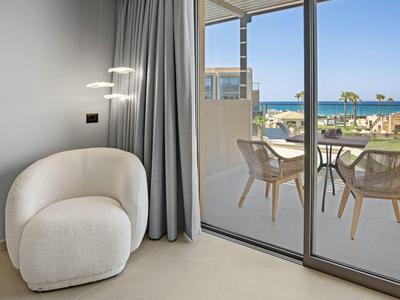 Heller Raum mit weißem Sessel, grauem Vorhang und Balkon mit Tisch, Stühlen und Meerblick.