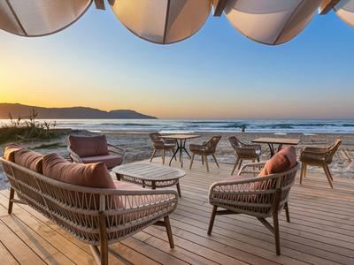 Sitzbereich mit Rattanmöbeln auf Holzterrasse am Meer bei Sonnenuntergang unter großen Sonnenschirmen.