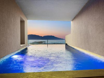 Schwimmbad mit blauer Beleuchtung, eingerahmt von Betonwänden, Blick auf Meer und Sonnenuntergang.