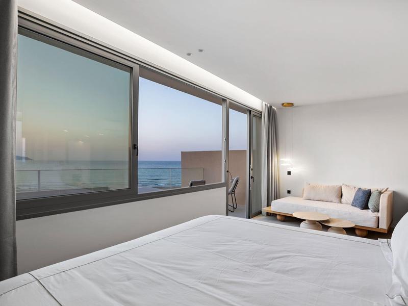 Helles Schlafzimmer mit großem Fenster, weißer Bettdecke, Sofa und Meerblick bei Sonnenuntergang.