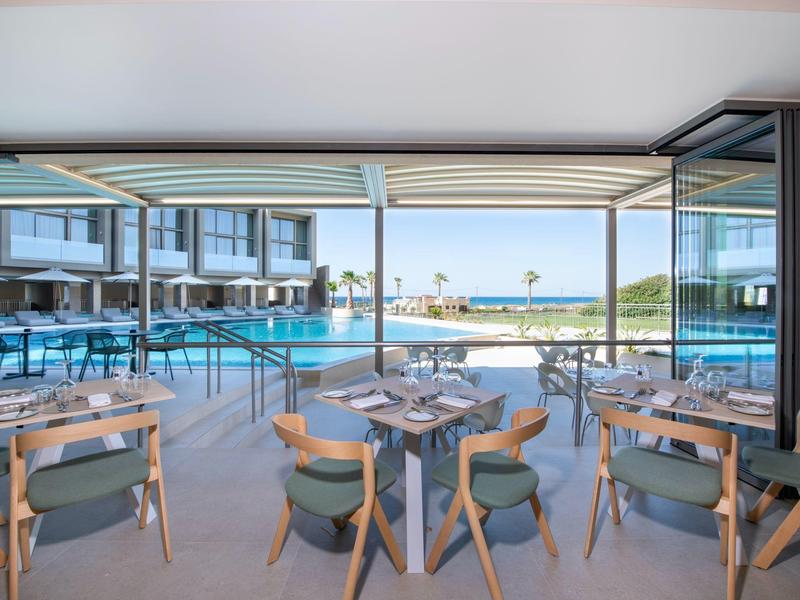 Moderner Essbereich mit Holztischen und Stühlen, Poolblick und offener Glasfront zum Meer.