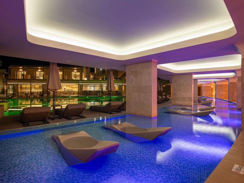 Moderner Innenpool mit blauer Beleuchtung, Liegen im Wasser und gemütlicher Sitzbereich dahinter.