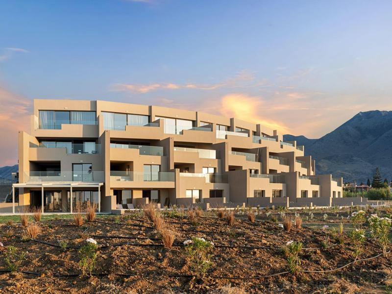 Modernes Wohnhaus mit Balkonen vor bergiger Landschaft und blauem Himmel bei Abendlicht.