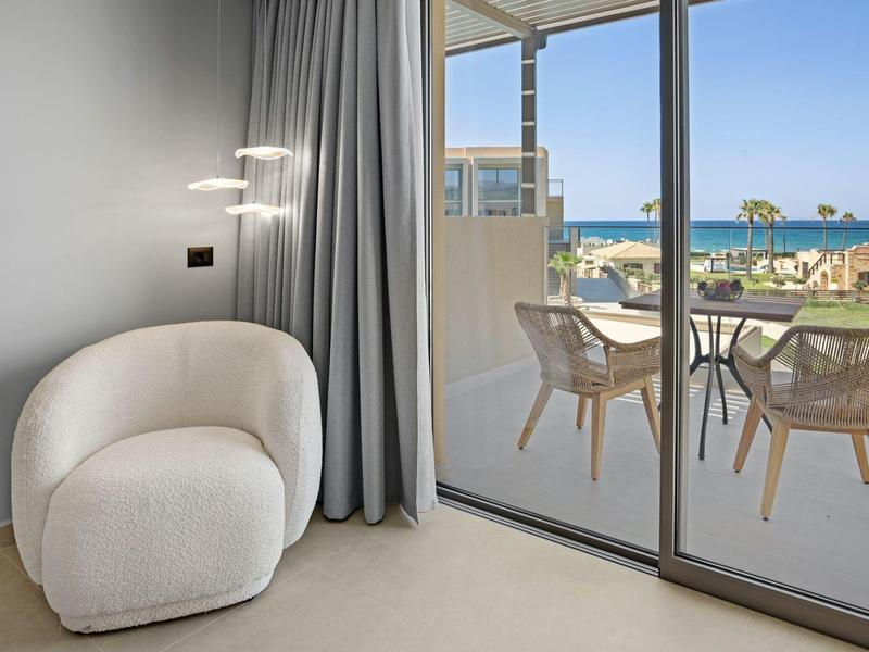 Heller Raum mit weißem Sessel, grauem Vorhang und Balkon mit Tisch, Stühlen und Meerblick.