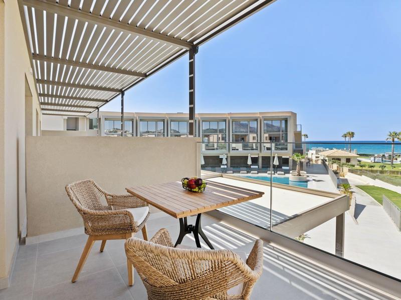 Balkon mit Holztisch, zwei Stühlen und Pergola, Blick auf Wohngebäude und Poollandschaft.