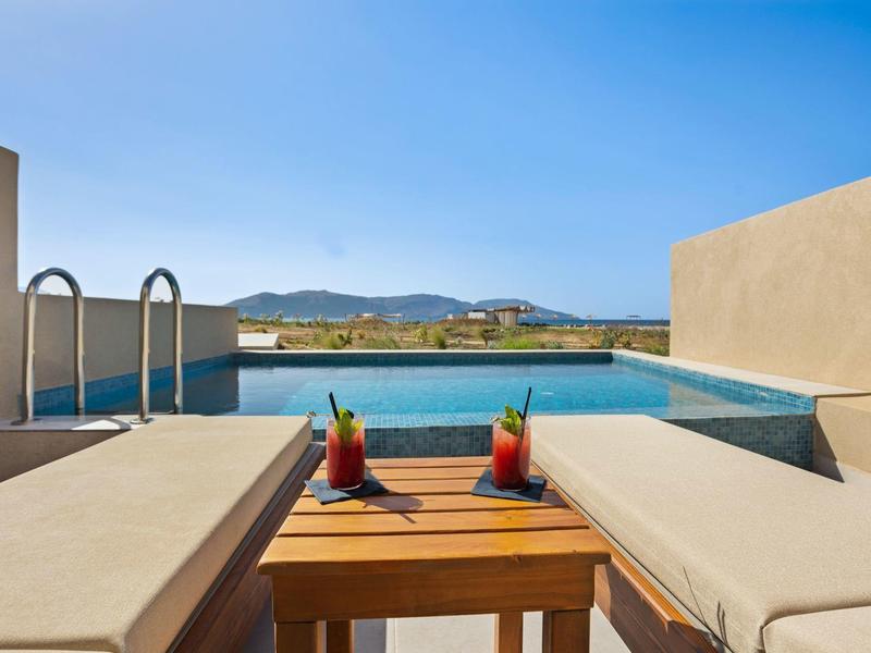 Zwei Liegen mit Cocktails am Holzttisch vor einem Pool, dahinter Berge und blauer Himmel.