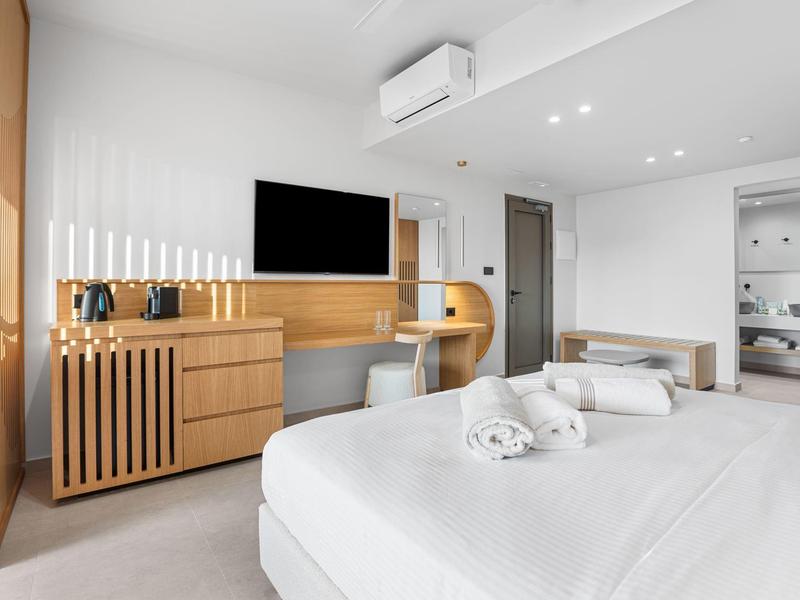 Helles modernes Hotelzimmer mit Doppelbett, hellem Holz, TV und offenem Badbereich.