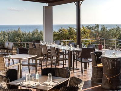 Terrassenrestaurant mit Holzboden, Tischen, Stühlen und Blick auf Meer und grüne Bäume am Horizont.