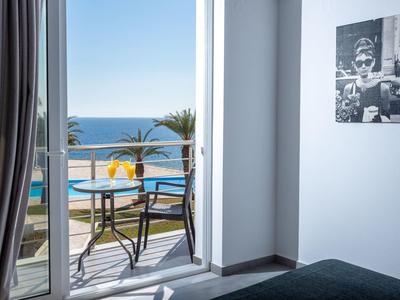 Vista desde una habitación de hotel con balcón hacia la piscina, el mar y palmeras en un día soleado.