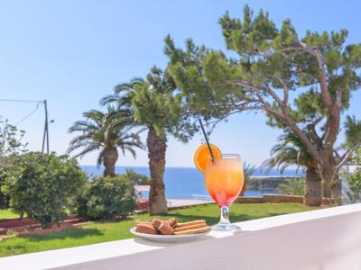 Bicchiere di cocktail e biscotti su un balcone con vista su giardino e mare sotto cielo limpido.
